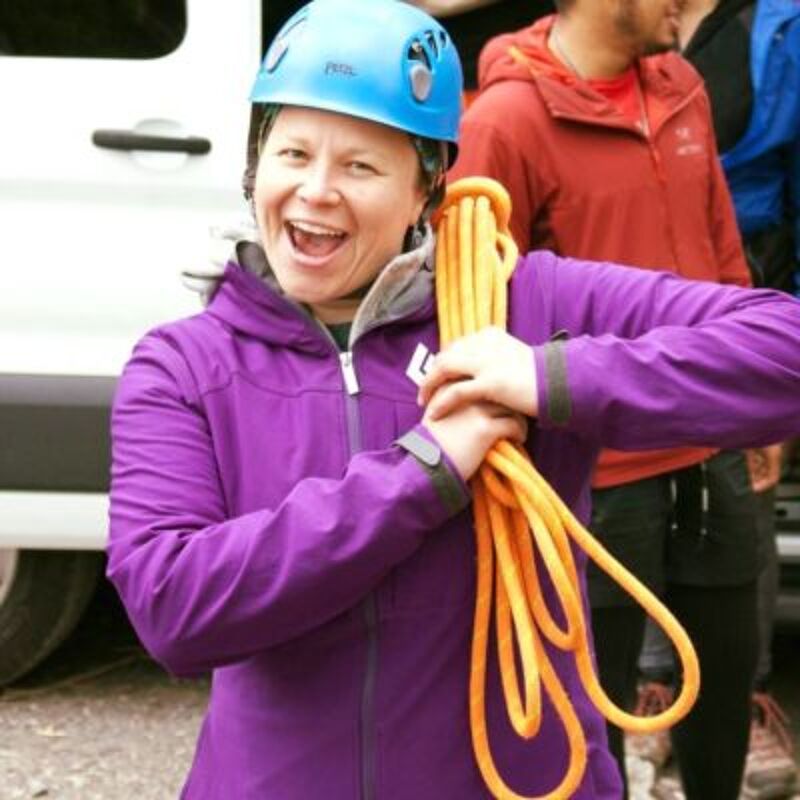 The image shows a woman wearing a blue helmet and a purple jacket, holding an orange rope. She is smiling and appears to be excited. In the background, there are other people, one wearing a red jacket. The setting seems to be outdoors, possibly before or after a climbing activity.